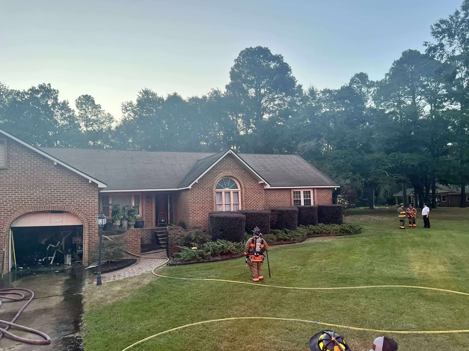 Two firefighters in gear stand on the front lawn of a brick house, with fire hoses stretched out across the grass and smoke in the background.