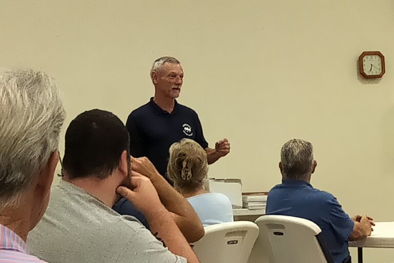 A man stands and speaks to a group of seated people in a classroom or meeting room setting.