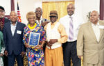 A group of five elderly people posing indoors, one woman holding a blue plaque, standing in front of a golden backdrop with an American flag visible.