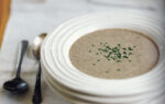 Creamy mushroom soup served in a white bowl, topped with chopped fresh herbs, placed on a white tablecloth beside two spoons.