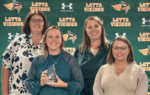 Four women smiling in front of a "Latta Vikings" backdrop; one woman is holding a triangular-shaped award.