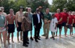 A group of people standing together by a swimming pool outdoors on a sunny day, dressed in various casual and formal outfits.