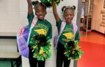Two young girls smiling, wearing green cheerleading shirts and holding green and yellow pom-poms, standing indoors with backpacks.