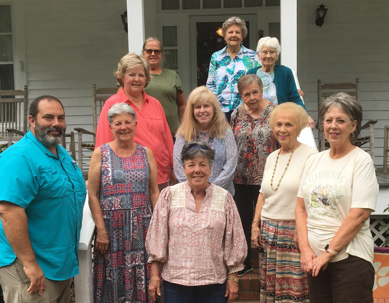 A group of older adults, both men and women, stand and smile together on the steps of a porch outside a white house.