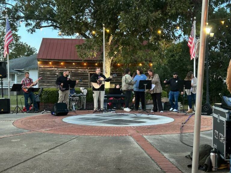 A group of musicians perform outdoors on a circular brick area, surrounded by trees and buildings, with string lights and an American flag visible.