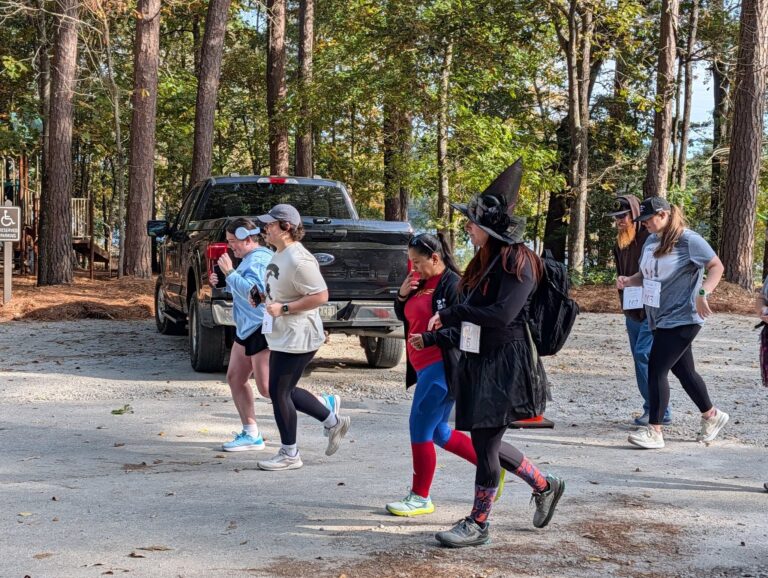 People in costumes, including Spider-Man and a witch, walk outdoors in a wooded area near a black pickup truck, participating in an event.