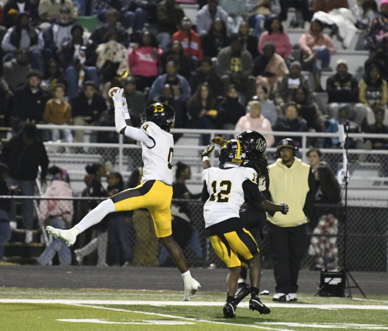 A football player in a white and yellow uniform jumps to catch the ball during a game, with spectators watching in the stands.