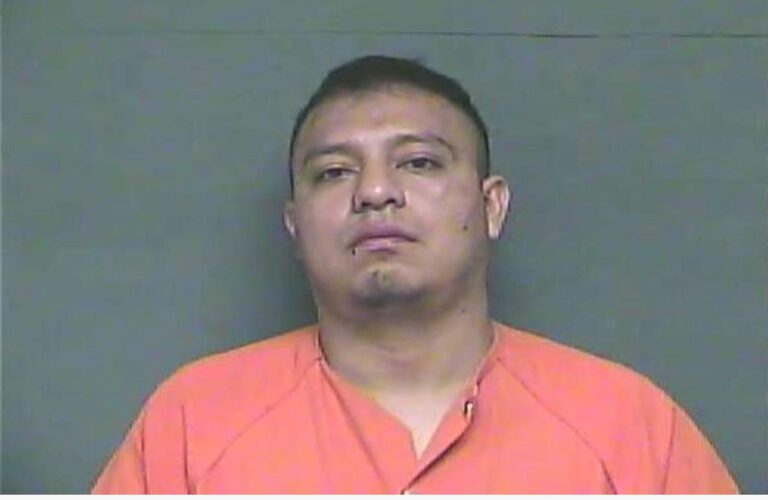 A man in an orange prison jumpsuit stands against a plain gray background, looking straight at the camera with a neutral expression.