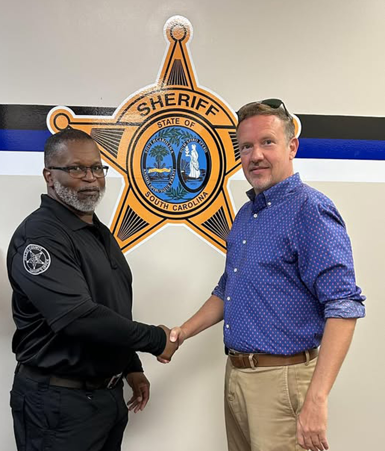 Two men shaking hands in front of a sheriff’s department emblem on a wall.
