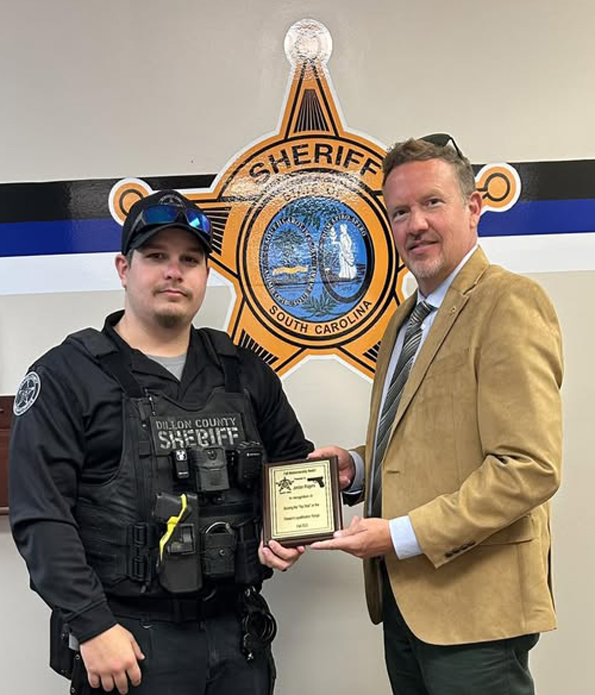 A sheriff's deputy in uniform receives a plaque from a man in a tan blazer, standing in front of a sheriff's office emblem.