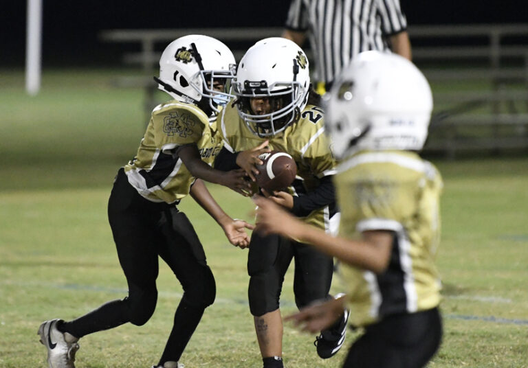 Three young football players in gold and black uniforms are playing during a game, with a referee visible in the background.