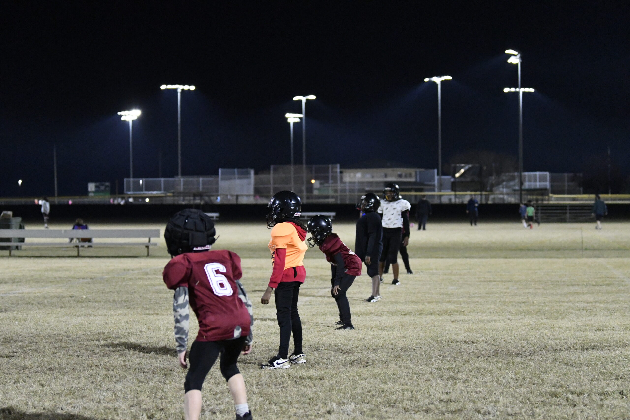 Kids in football gear line up on a field at night under bright stadium lights, preparing for a play or practice session.