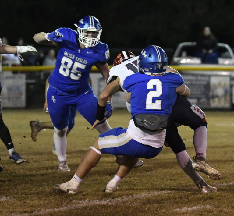 Two football players in blue uniforms tackle an opposing player in black during a nighttime game on a grass field.