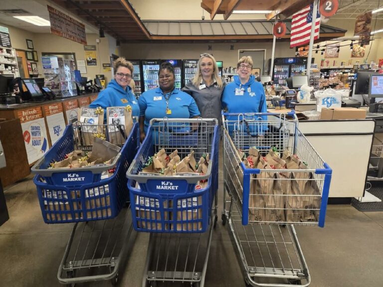 Four people in blue shirts stand behind three shopping carts filled with paper bags inside a grocery store, with an American flag displayed in the background.