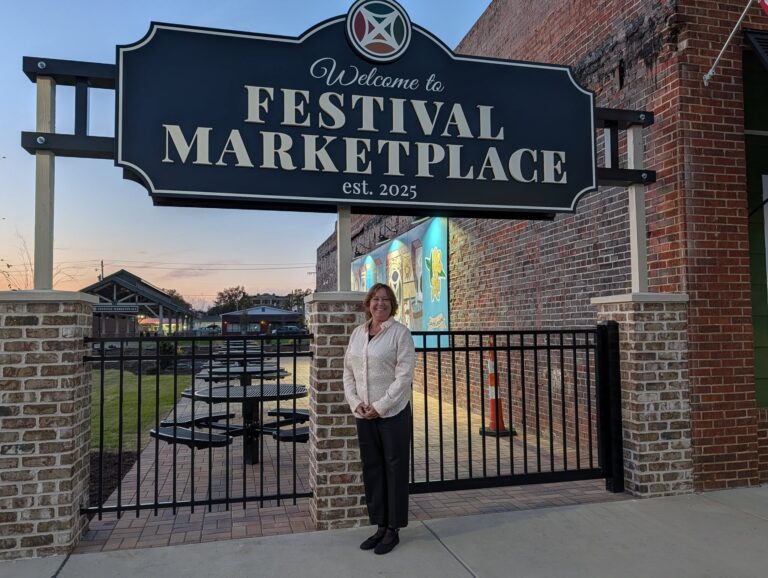 A person standing under a large "Festival Marketplace" sign at an outdoor venue with picnic tables and a mural visible in the background.