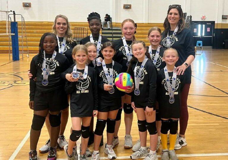 A girls' volleyball team poses indoors, wearing medals and matching uniforms, with two adult coaches standing behind them. One girl holds a colorful volleyball.