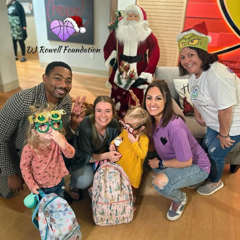 Group posing with Santa Claus; two children with backpacks and festive glasses, three adults smiling, and another woman in a Christmas hat. Indoors, holiday decorations visible.