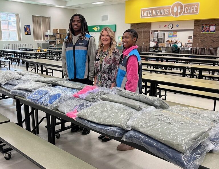 Three people stand behind tables covered with neatly folded jackets inside a cafeteria or dining hall.