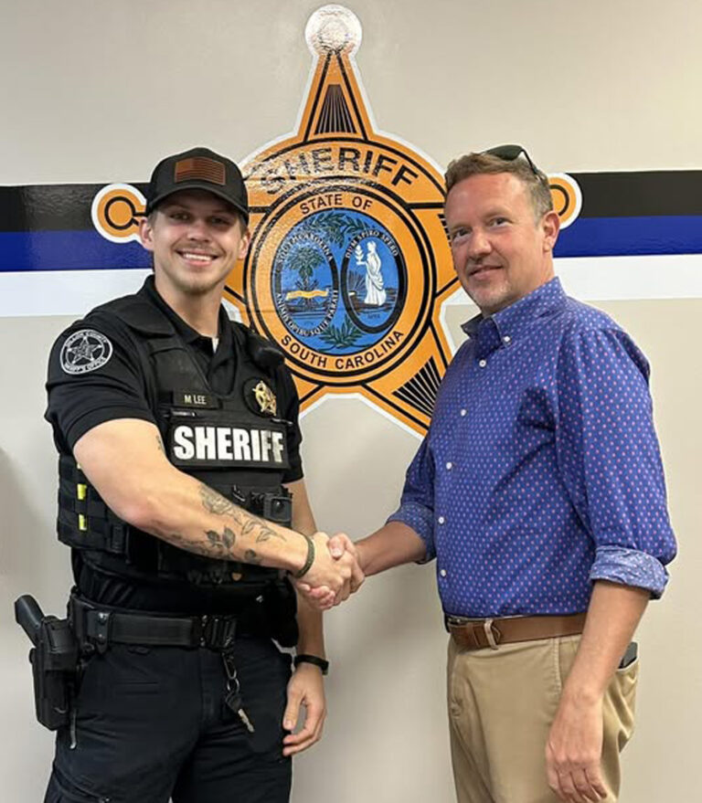 A sheriff and another man shake hands in front of a wall with a sheriff's badge emblem.