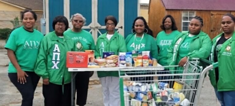Five women in green shirts stand behind a table and shopping cart filled with food donations, outdoors in front of blue and brown structures.