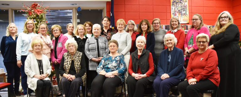 A group of older women posing together indoors, with some sitting in chairs and others standing behind them, against a red and tan wall background.