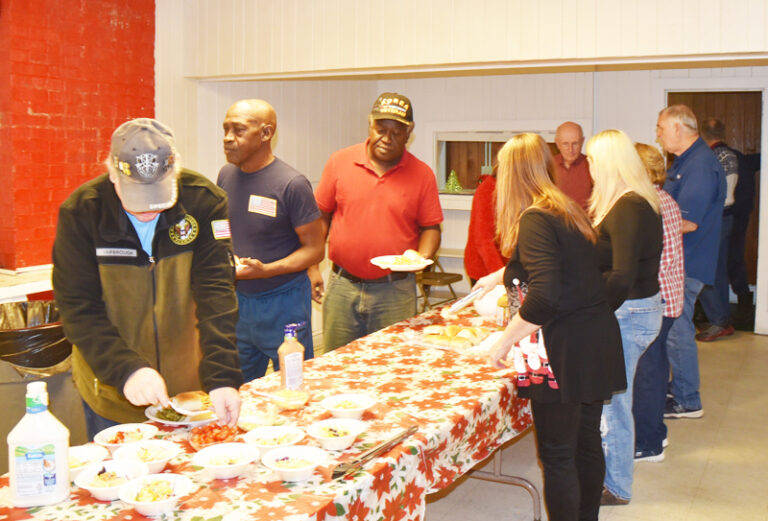 People standing and serving themselves food from a buffet table covered with a colorful tablecloth in a well-lit room.