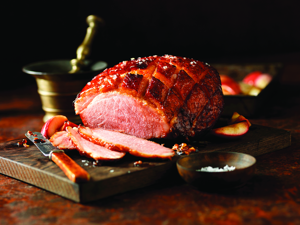 A roasted ham with a glazed, crisscrossed surface is sliced on a wooden cutting board, surrounded by apples and a knife.