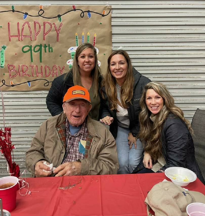 Older man in an orange cap sits at a table with three smiling women behind him, celebrating his 99th birthday with decorations in the background.