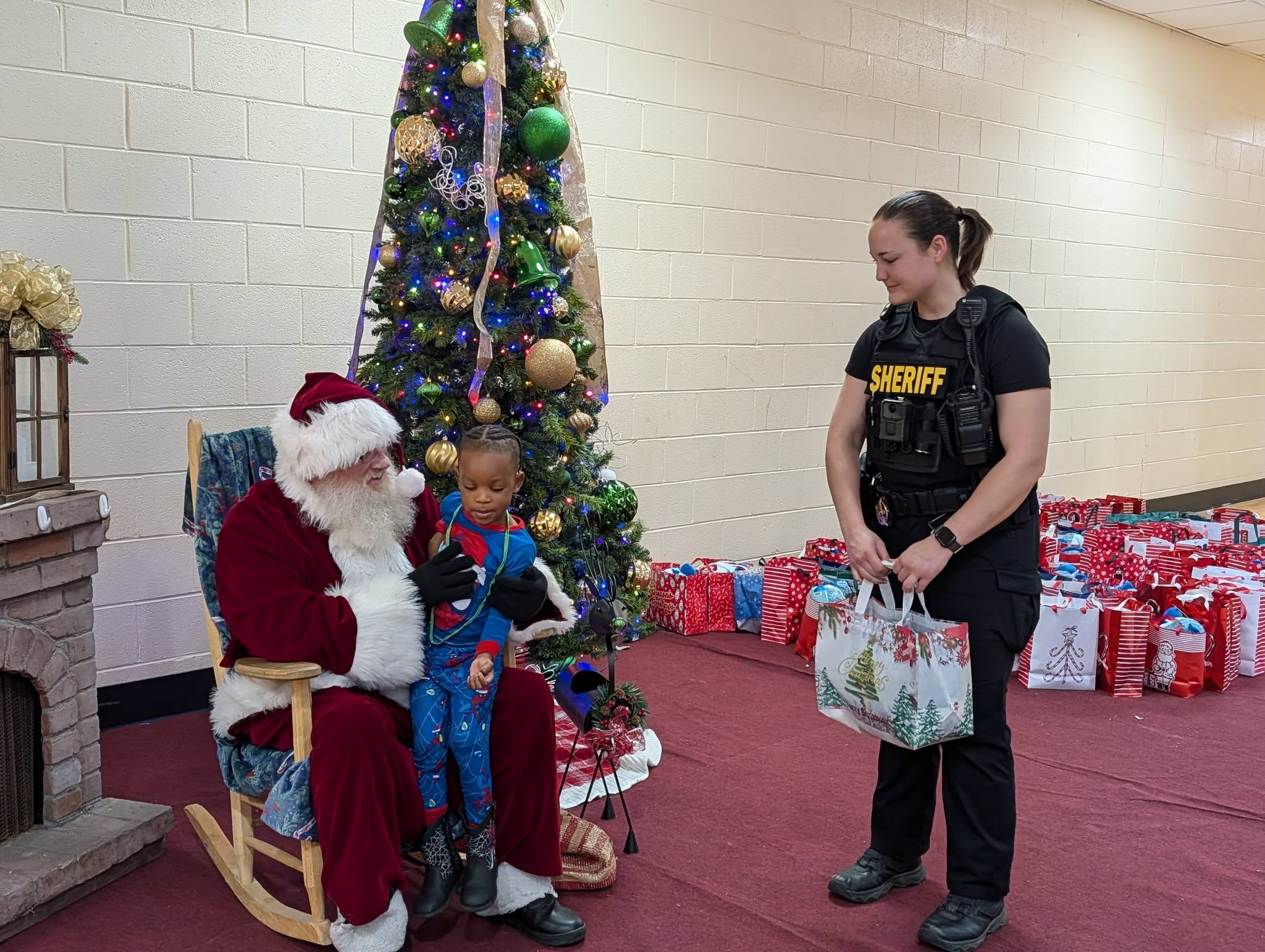 A child sits on Santa's lap near a decorated Christmas tree, while a woman in a sheriff shirt stands nearby holding a gift bag.