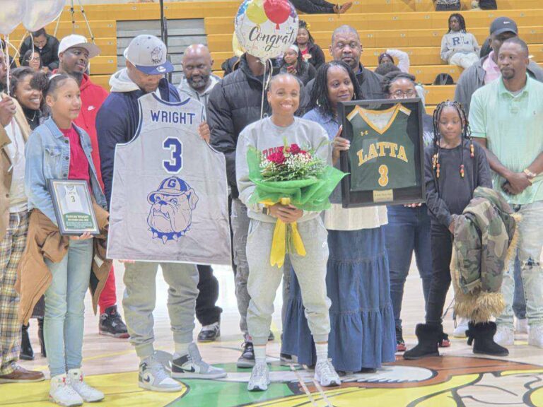 A group of people stands in a gymnasium; a young woman in the center holds flowers, others display sports jerseys.