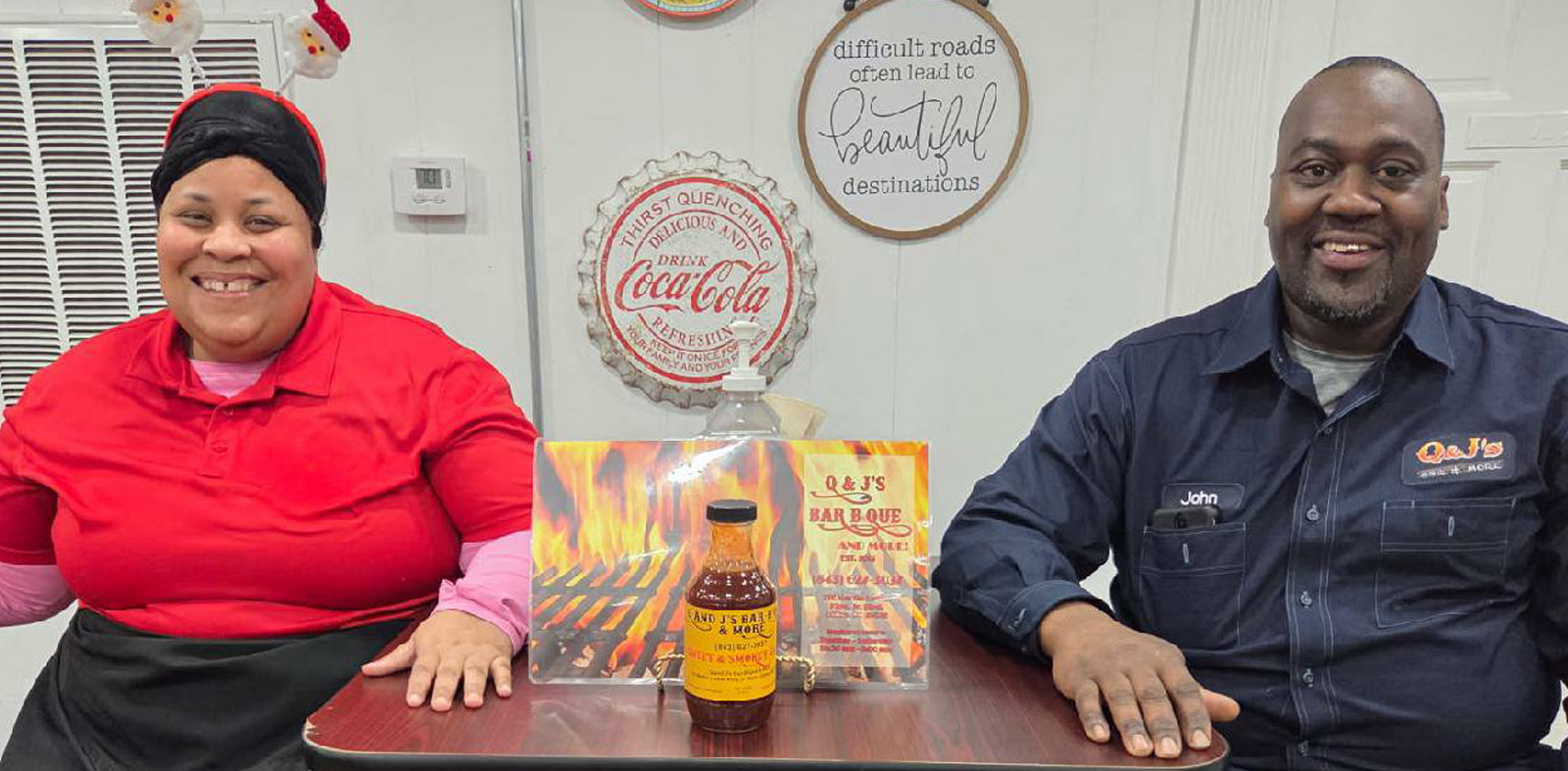 Two people sit at a table with a bottle of barbecue sauce and a barbecue-themed display, Coca-Cola signs hang on the wall behind them.