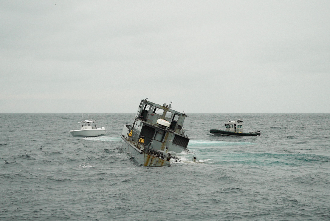 A partially submerged ship tilts in rough waters, with two smaller boats nearby under a cloudy sky.