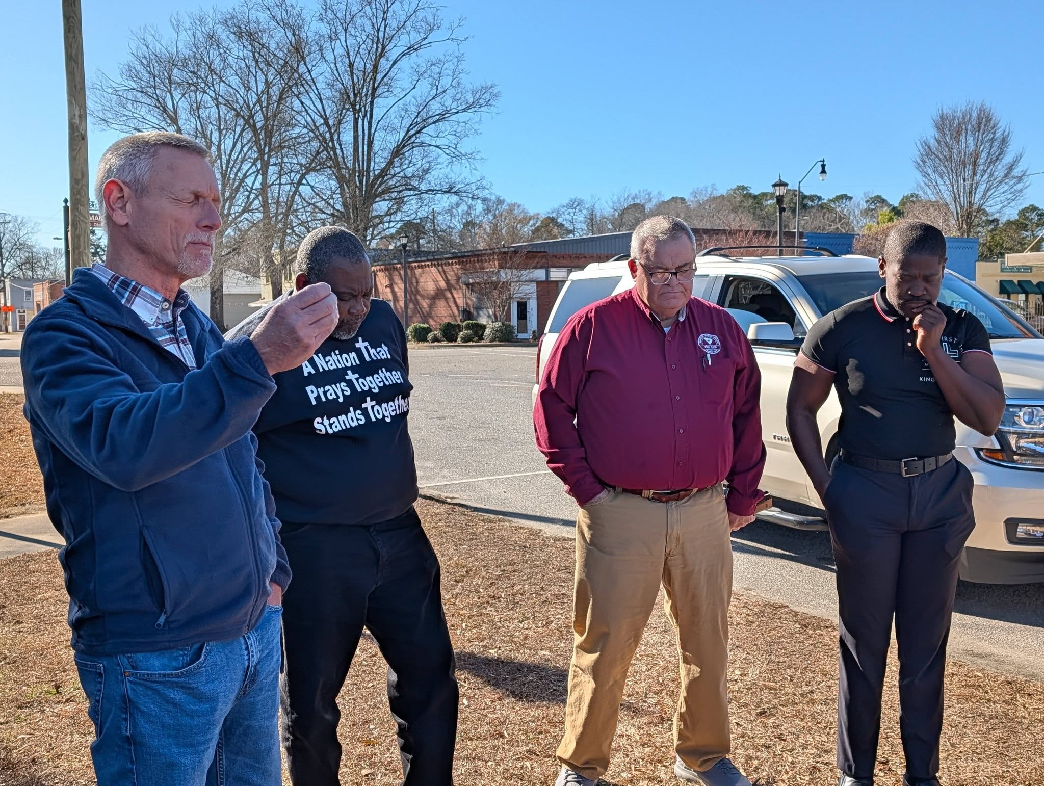 Four men standing outdoors in conversation near parked vehicles on a sunny day, trees and buildings visible in the background.