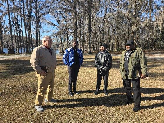 Four people standing outside on grass, wearing jackets and coats, with trees and a blue sky in the background.