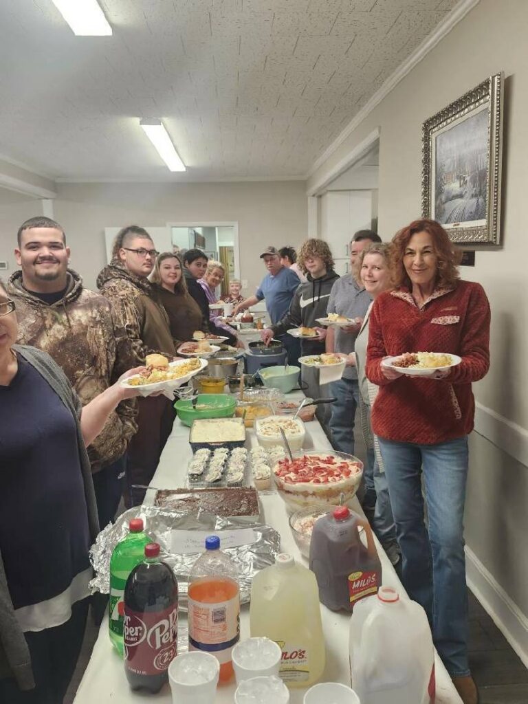 A group of people stand in line at a buffet table filled with various foods and drinks, smiling and holding plates in a bright indoor room.