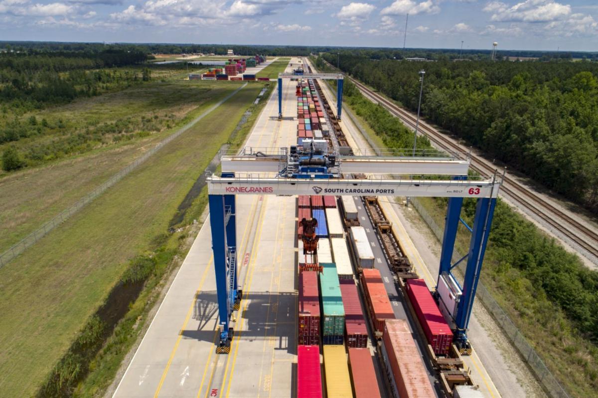 Aerial view of a railway yard with colorful shipping containers and large blue cranes, surrounded by greenery and tracks under a partly cloudy sky.