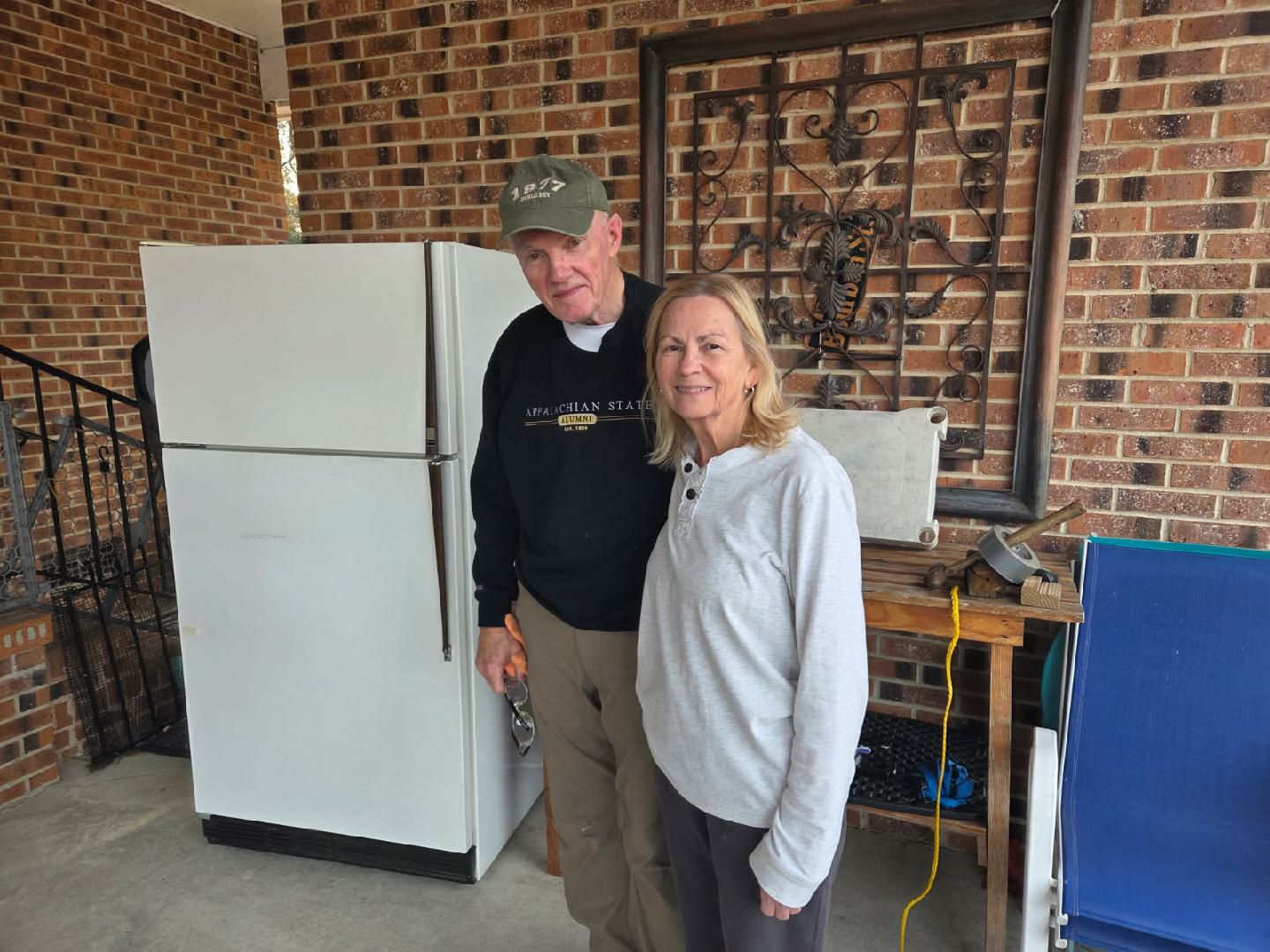 older man and woman stand together indoors near a refrigerator, smiling at the camera. There is brick wall and metal decor in the background.
