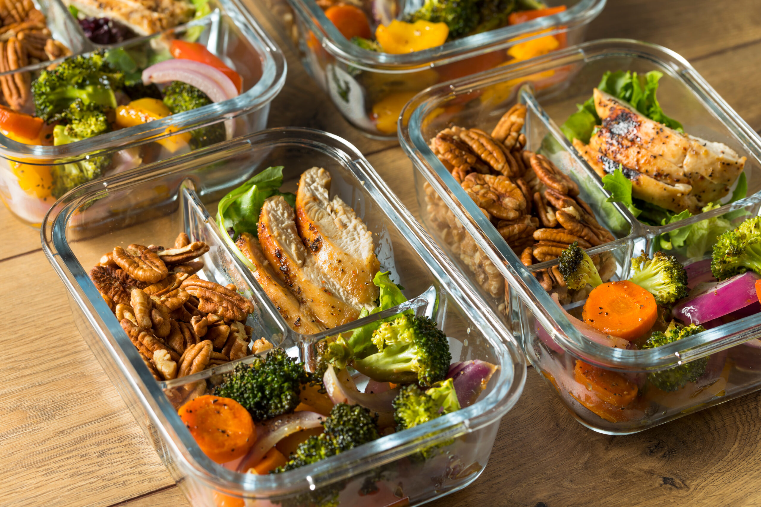 Glass meal prep containers filled with grilled chicken, roasted vegetables like broccoli and carrots, and granola clusters, arranged neatly on a wooden surface.