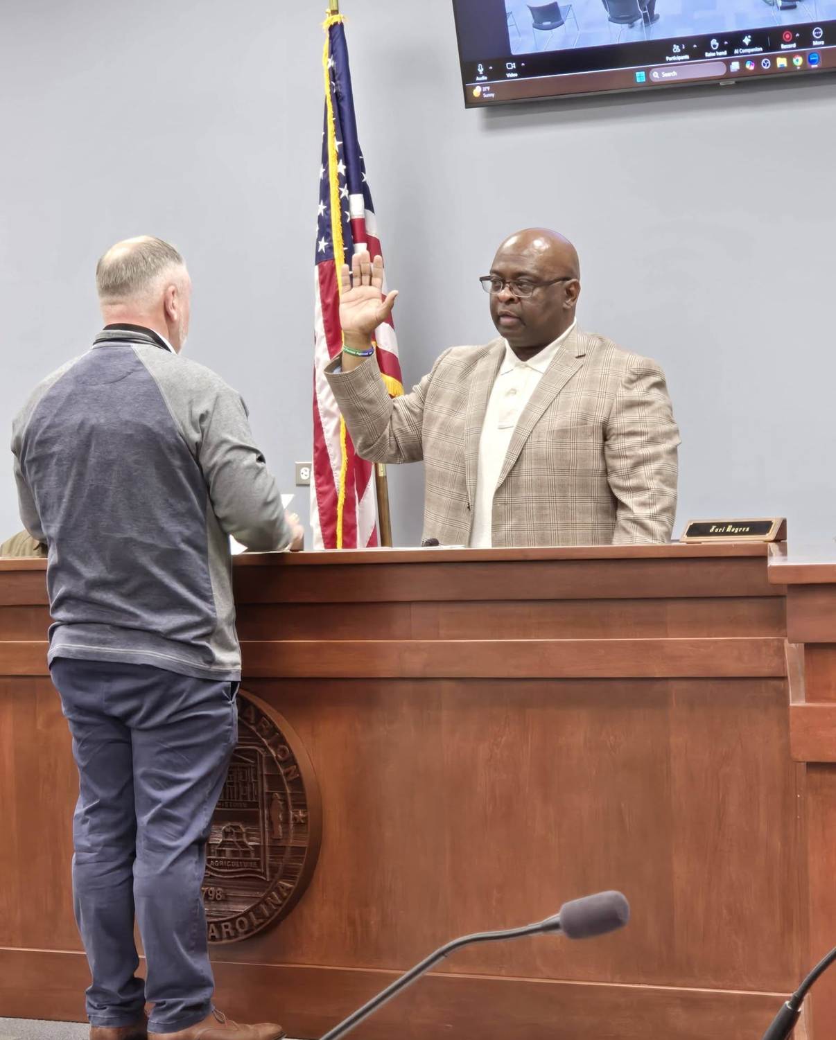 A man stands in front of a judge taking an oath in a courtroom, with an American flag in the background.