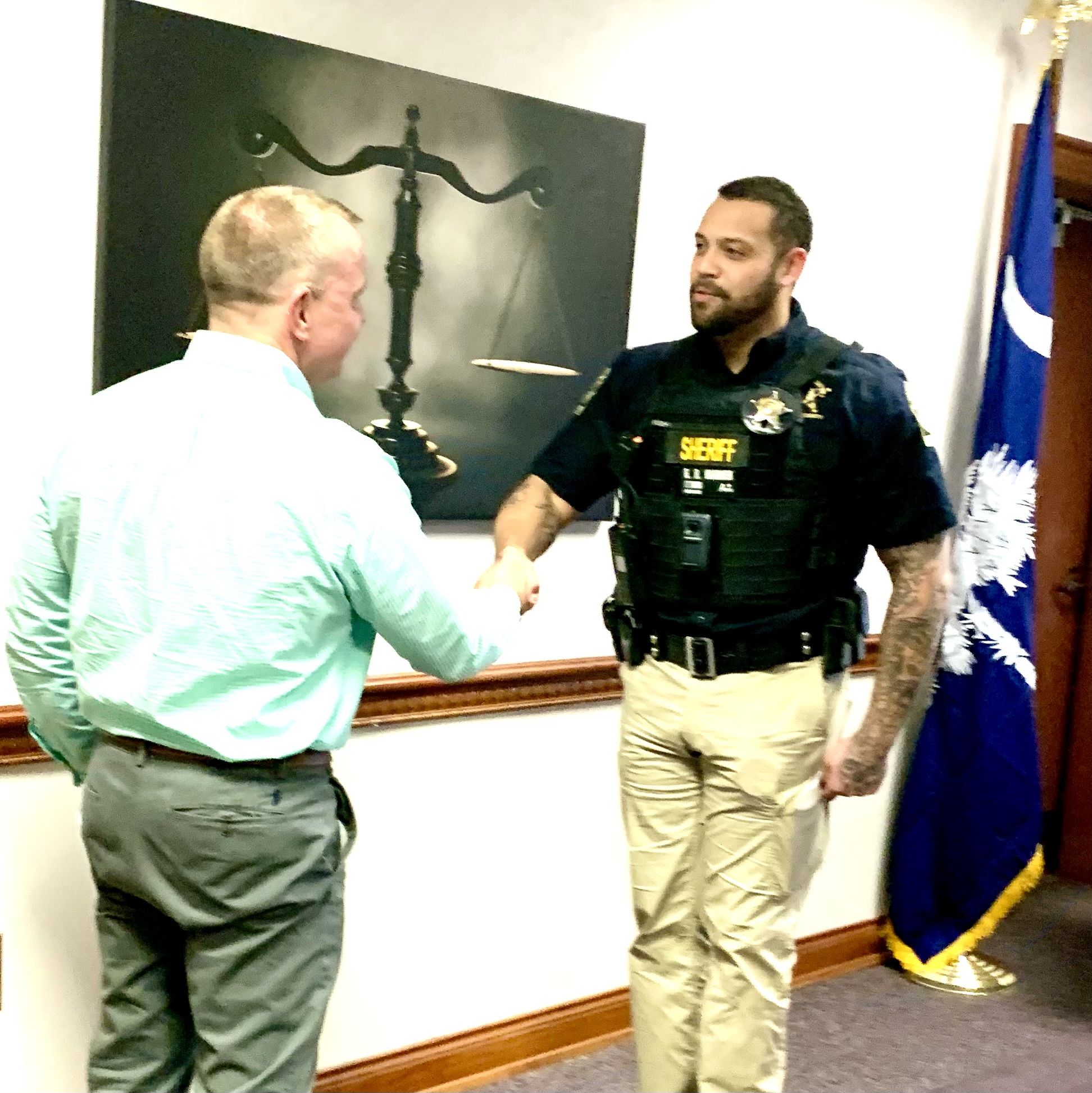 A police officer and a man in business attire shake hands in an office, with a scales of justice picture and a flag in the background.