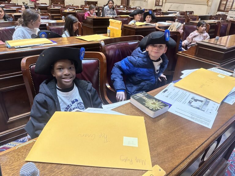 Children in a classroom wear colonial-style hats, sitting at desks with yellow folders and papers, smiling and participating in an educational activity.