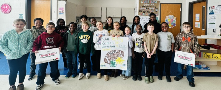 A group of children standing together in a classroom, smiling and holding a colorful handmade sign with drawings and text about sports.