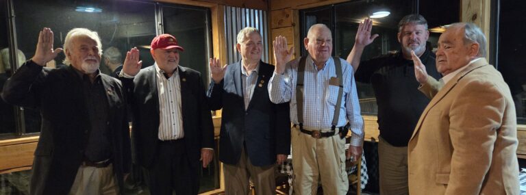 Four older men standing indoors, raising their right hands, dressed in business casual and formal attire, smiling, posing for a photo in a warmly lit room.
