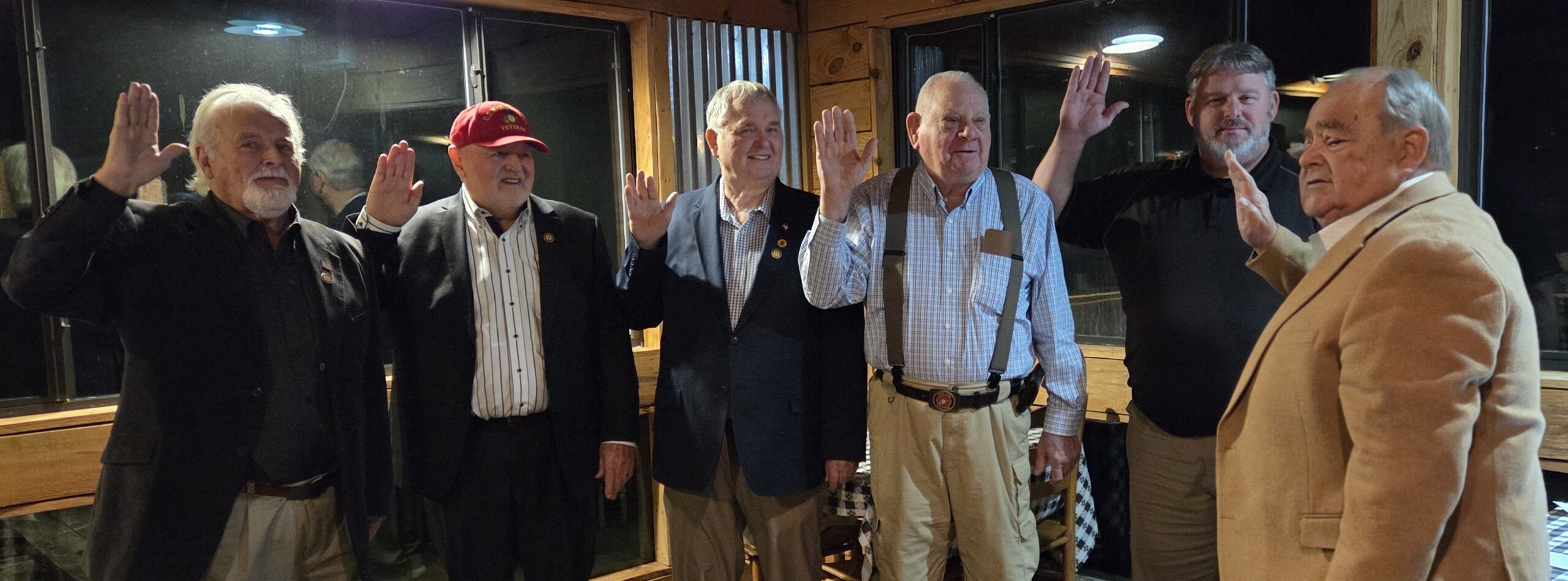 Four older men standing indoors, raising their right hands, dressed in business casual and formal attire, smiling, posing for a photo in a warmly lit room.