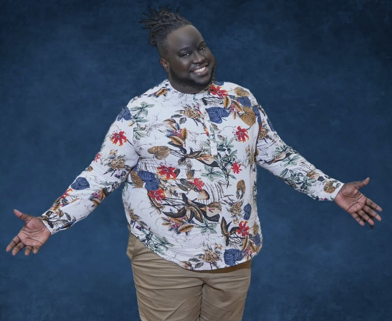 A smiling person in a floral shirt and tan pants stands with arms open against a blue textured background.