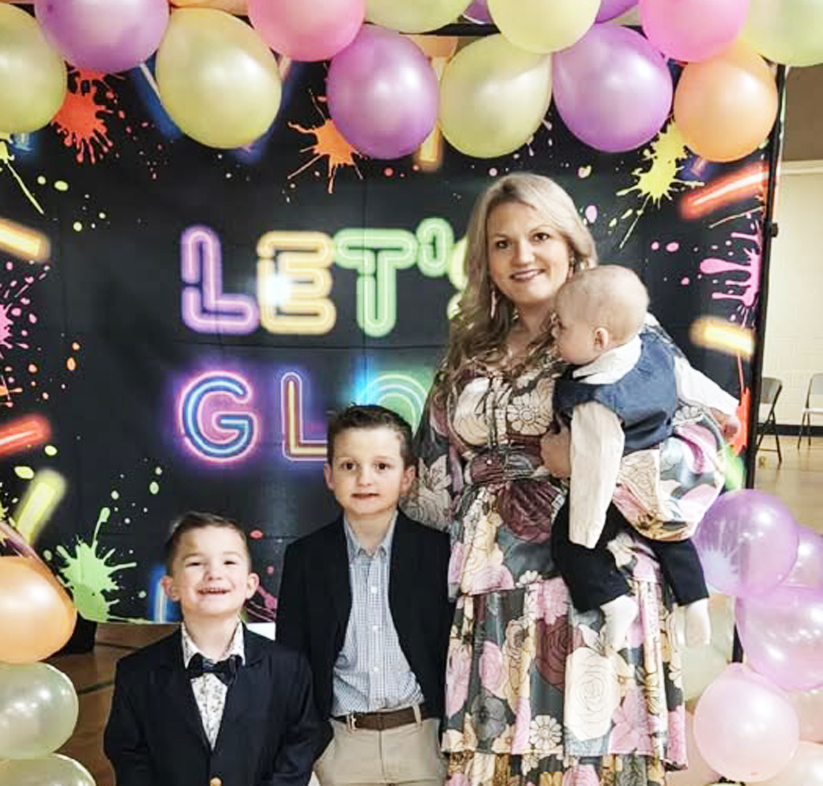 A woman holding a baby stands with two young boys in front of a colorful "LET'S GLO" sign and pastel balloon decorations.
