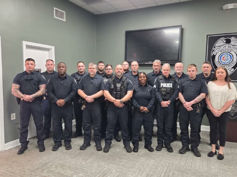 A group of police officers in uniform stand together in a room with two large TVs on the wall behind them.