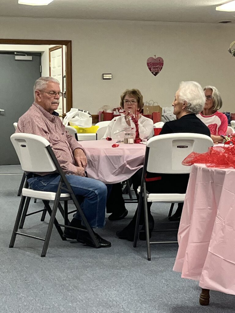 Four older adults sit around a table covered with a pink tablecloth in a decorated room, conversing and enjoying a gathering.