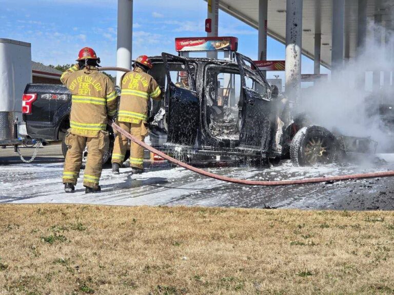 Firefighters extinguish a burning vehicle near a gas station, with smoke rising from the car.