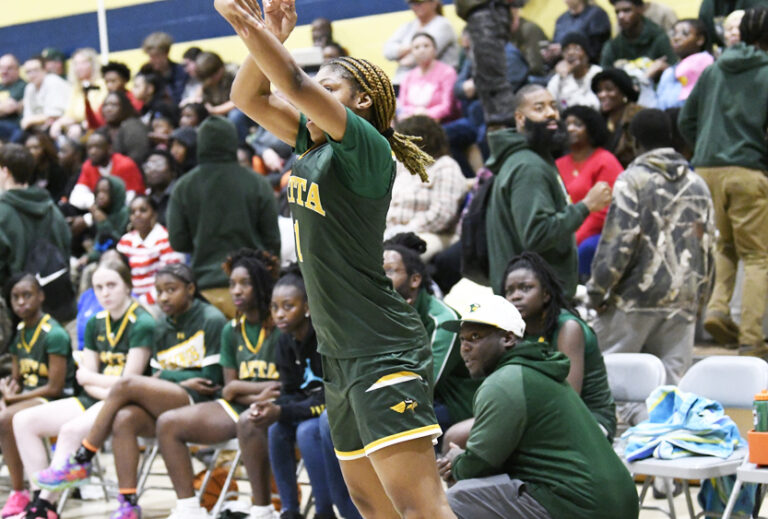 A female basketball player in a green uniform takes a jump shot during a game, with teammates and spectators watching from the sidelines.